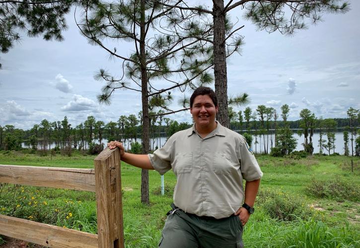 Park Ranger Sam Louzan smiling for the camera with pine trees and a lake behind him