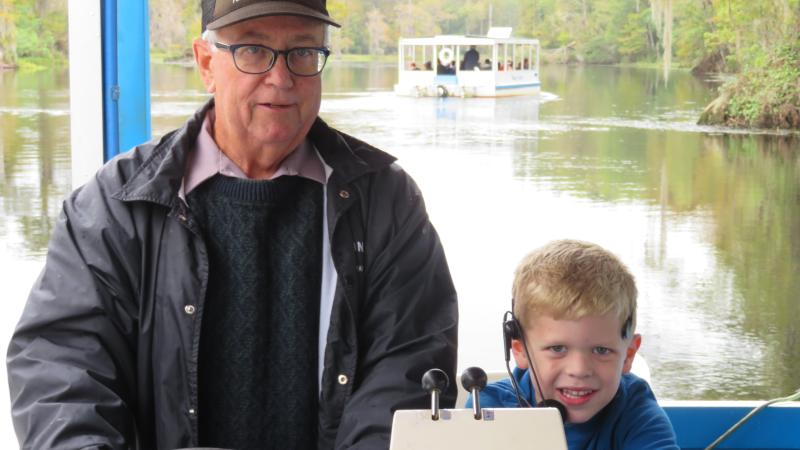 Volunteer at captains wheel of tour boat with young boy sitting nearby as co-captain. River boats can be seen on the river in the distance.